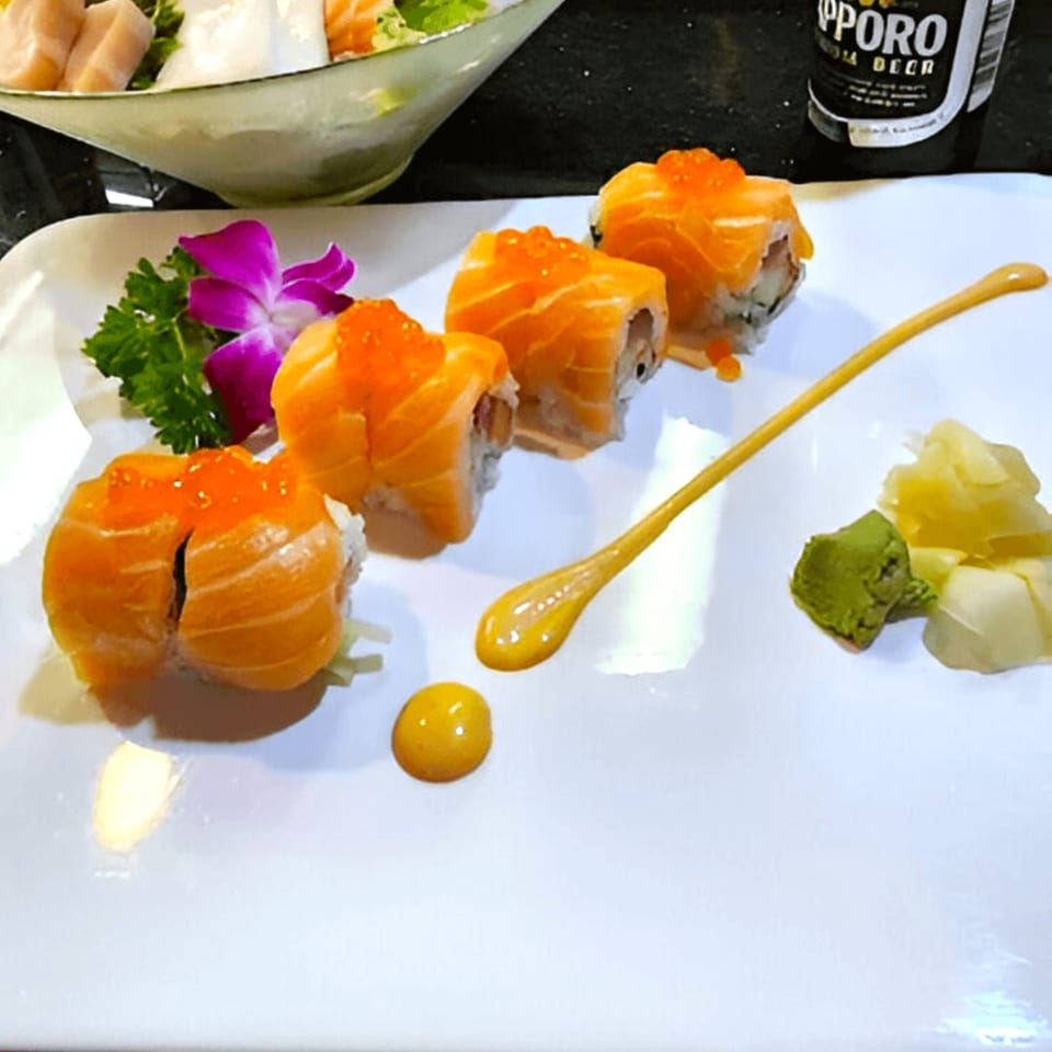 Four salmon sushi rolls with roe, wasabi, ginger, and a decorative sauce swirl on a white plate.